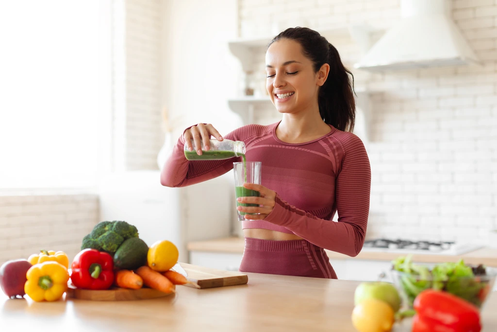 A side view of Isabella "Izzy" Rossi pouring a vibrant green smoothie into a glass, surrounded by fresh fruits and vegetables on her kitchen counter.