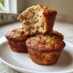 A stack of three golden-brown banana protein muffins, one broken open to show the moist, speckled interior texture.