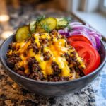 Close-up of a cheeseburger bowl featuring seasoned ground beef topped with melted cheddar cheese, served with pickles, tomato, and red onion slices.
