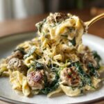 A spoonful of creamy chicken sausage pasta being lifted, showing melted cheese strings and chunks of sausage and spinach.