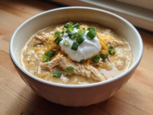 A close-up of a bowl of creamy crockpot white chicken chili topped with shredded cheese, sour cream, and green onions.