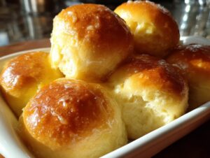 Close-up of several golden-brown, fluffy dinner rolls stacked in a white dish, glistening with a light glaze.