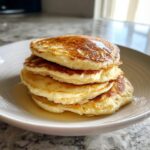 A tall stack of four fluffy greek yogurt pancakes drizzled with syrup, served on a white plate.