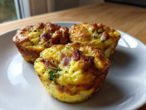 Three perfectly baked Egg Muffin Cups featuring visible chunks of ham and green broccoli florets on a white plate.