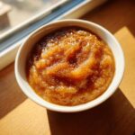 A close-up of a white bowl filled with rich, brown homemade applesauce, sprinkled with cinnamon, sitting in sunlight.