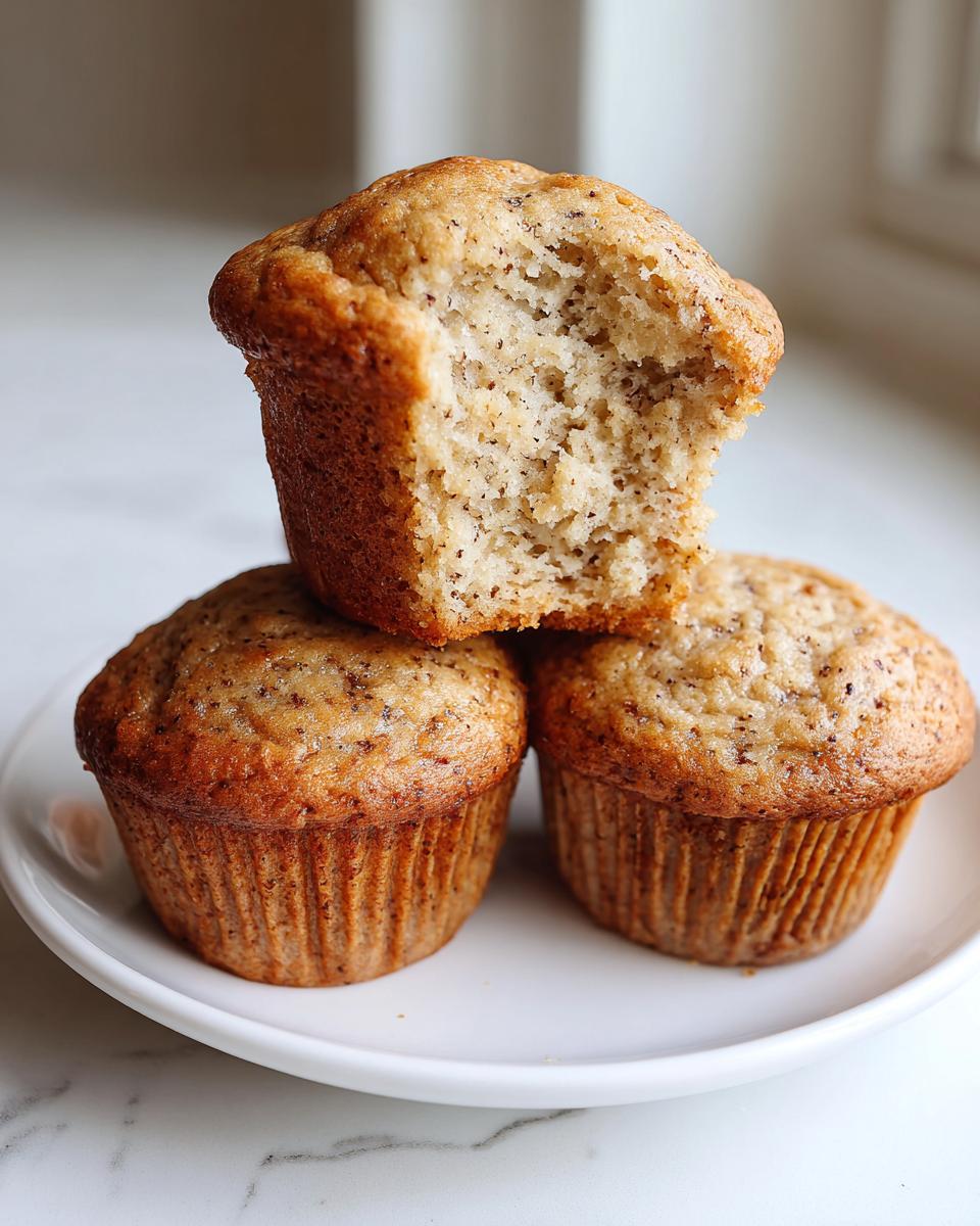 Three banana protein muffins stacked on a white plate, with the top muffin having a bite taken out revealing the moist interior.
