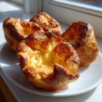 Four golden brown, puffed-up yorkshire pudding items sitting on a white plate near a sunlit window.