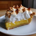 A close-up of a tall slice of lemon meringue pie showing bright yellow filling and toasted meringue peaks.