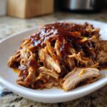 Close-up of tender, shredded slow cooker pulled pork smothered in rich barbecue sauce on a white plate.