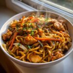 Close-up of a steaming bowl filled with delicious chicken chow mein recipe, featuring noodles, chicken pieces, carrots, and cabbage.