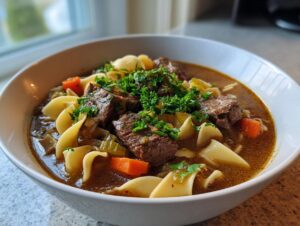 A close-up of a white bowl filled with rich beef noodle soup, featuring chunks of beef, egg noodles, carrots, and fresh parsley.