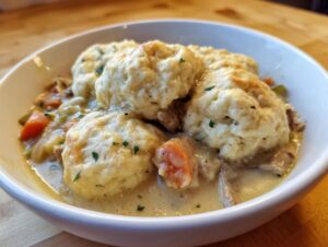 A close-up of a white bowl filled with creamy chicken and dumpling stew, topped with fluffy dumplings.
