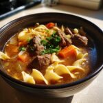 Close-up of a dark bowl filled with rich broth, tender chunks of beef, egg noodles, carrots, and parsley, showcasing the beef noodle soup.