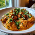 Close-up of rich, orange-hued butter chicken pieces generously topped with fresh cilantro in a white bowl.