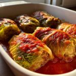 Close-up of homemade cabbage rolls stuffed with meat, simmering in a rich tomato sauce in a white baking dish.