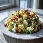 A mound of creamy pea salad mixed with chopped eggs and topped generously with crispy bacon bits, served on a white plate.