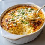 A spoonful being lifted from a hot, baked street corn dip topped with crumbled white cheese and cilantro.