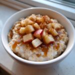A white bowl filled with creamy apple cinnamon oatmeal, topped with diced red apples, cinnamon, and caramel drizzle.