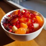 Close-up of a bright winter fruit salad in a white bowl, featuring vibrant orange segments and deep red pomegranate seeds.