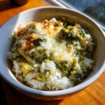Close-up of a serving of cheesy broccoli rice casserole in a white bowl, featuring melted, slightly browned cheese over white rice and green broccoli florets.