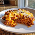 A fork lifts a bite of cheesy mexican rice casserole mixed with seasoned ground beef from a white plate.