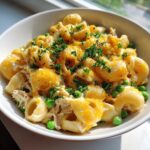 A close-up of creamy, cheesy tuna pasta mixed with bright green peas, topped with melted cheddar and fresh parsley.