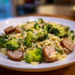 A close-up of a white bowl filled with chicken sausage and broccoli orzo, featuring browned sausage slices and bright green florets.