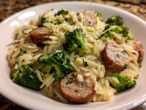 A close-up of creamy chicken sausage and broccoli orzo served in a white bowl, featuring browned sausage slices and bright green florets.