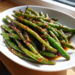 A white bowl filled with vibrant, blistered chinese green beans with garlic sauce glistening under natural light.