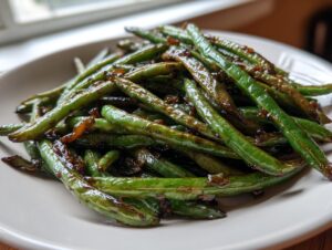 A close-up of vibrant, glossy chinese green beans with garlic sauce piled on a light-colored plate.