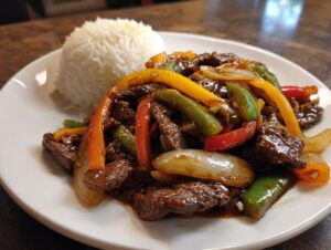 A plate of glossy Chinese pepper steak stir-fry with colorful bell peppers and onions, served alongside a mound of white rice.