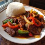 A close-up of a plate featuring savory Chinese pepper steak mixed with colorful bell peppers and onions, served next to a mound of white rice.
