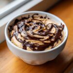 A creamy peanut butter dip topped with a rich swirl of melted dark chocolate drizzle in a white bowl.