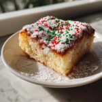 A square slice of a rich, gooey dessert, resembling Christmas gooey butter cookies, topped with powdered sugar and red and green sprinkles.