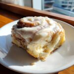 A single, fluffy cinnamon roll generously topped with white icing, sitting on a white plate near a sunlit window.