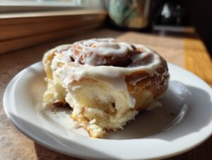 A single, fluffy cinnamon roll with thick white icing resting on a white plate, showcasing the soft interior.
