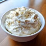 A close-up of rich, creamy clam dip swirled high in a white bowl, lightly garnished with spices.
