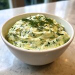 Close-up of a creamy, vibrant green dill dip garnished with fresh dill sprigs in a white bowl.