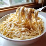 A fork lifting strands of fettuccine coated in thick, creamy homemade alfredo sauce from a white bowl.