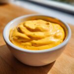Close-up of thick, bright orange nacho cheese sauce with visible spices in a small white bowl.