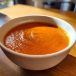 A close-up of a white bowl filled with vibrant orange tomato bisque, topped with cracked black pepper.