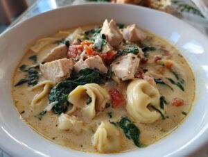 A close-up, mouthwatering bowl of creamy tortellini soup featuring chunks of chicken, wilted spinach, and diced tomatoes.