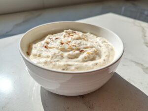 A close-up of creamy white pizza sauce speckled with spices, served in a white bowl on a marble counter.