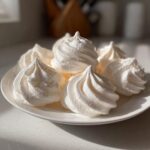 A close-up of several perfectly piped, crisp white meringue cookies stacked on a white plate.
