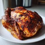 A perfectly cooked, golden-brown air fryer whole chicken with crispy, seasoned skin resting on a white plate.