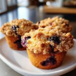 Three golden brown blueberry muffins topped with a generous layer of streusel crumb topping, sitting on a white plate.