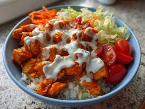 A vibrant buffalo chicken bowl featuring orange-glazed chicken pieces over rice, topped with ranch dressing, lettuce, tomatoes, and carrots.