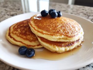 Close-up of a stack of fluffy almond flour pancakes drizzled with syrup and topped with fresh blueberries.