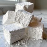 A pile of large, fluffy homemade marshmallows dusted generously with powdered sugar on a marble surface.