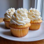 Close-up of four moist vanilla cupcakes topped with high swirls of creamy white vanilla frosting on a white plate.